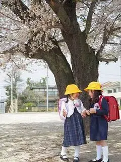 写真：校庭の桜の木の下で向かい合って笑顔でおしゃべりしている二人のランドセルを背負った子